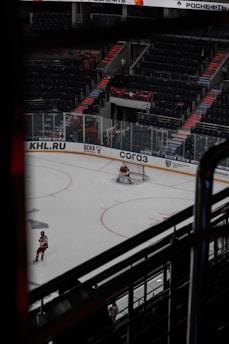 A hockey game taking place in an indoor arena with visible advertisements around the rink. The ice surface shows a goalie positioned in front of the net, and another player is near the boards wearing a red and white uniform. The arena seating is mostly empty, and there's a noticeable architectural element with steps leading up the stands.