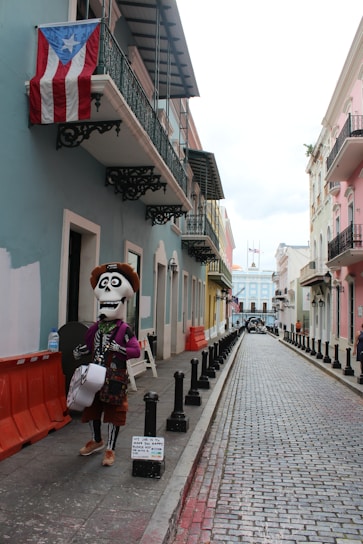 A narrow, colorful street with cobblestone pavement lined by buildings in pastel shades. A large puppet-like figure dressed in festive attire, resembling a skeleton with a hat and a guitar, stands on the sidewalk. An American flag and a Puerto Rican flag are visible hanging from a balcony.