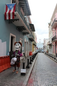A narrow, colorful street with cobblestone pavement lined by buildings in pastel shades. A large puppet-like figure dressed in festive attire, resembling a skeleton with a hat and a guitar, stands on the sidewalk. An American flag and a Puerto Rican flag are visible hanging from a balcony.
