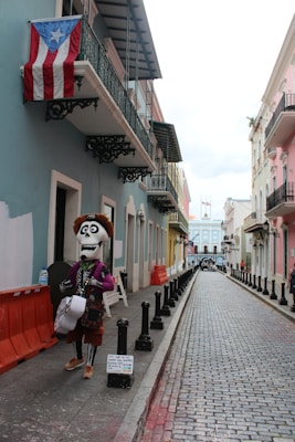 A narrow, colorful street with cobblestone pavement lined by buildings in pastel shades. A large puppet-like figure dressed in festive attire, resembling a skeleton with a hat and a guitar, stands on the sidewalk. An American flag and a Puerto Rican flag are visible hanging from a balcony.