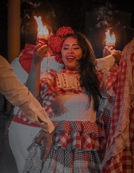 A woman dressed in a traditional outfit with a red and white checkered pattern is holding a lit candle. Her hair is adorned with flowers, and she is surrounded by people in similar attire, engaging in a celebratory or cultural event. The scene appears to be lively and festive.