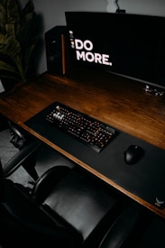 A well-organized computer desk setup featuring a large monitor displaying the words 'DO MORE.', a mechanical keyboard, a mouse on a mouse pad, a chair, and a speaker. A plant is positioned to the side, enhancing the aesthetic of the workspace.