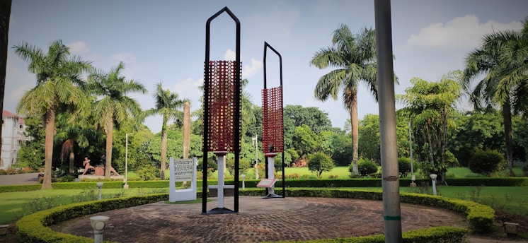 A park setting with tall palm trees and a circular arrangement of hedges. In the center, two tall, modern art sculptures made of vertical black frames and red patterns. The sky is partly cloudy, with some blue visible, adding to the serene ambiance of the greenery around.