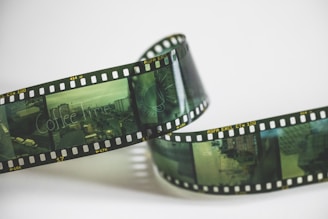 A strip of photographic film curling against a plain background. The film has visible frames showing various scenes, including buildings and a street, with one frame displaying the text 'Coffee Time'. The film strip has a green tint and contains numbers and text along its edges.