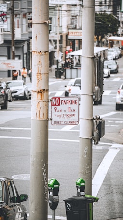 Urban street scene with a focus on a 'No Parking' sign for scheduled street cleaning. The background shows a busy street with cars and buildings, including a dry cleaner's. A weathered lamppost and green parking meters are in the foreground.