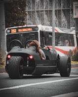 A sleek, black three-wheeled vehicle with red taillights is stopped at an urban intersection. A person wearing a furry coat is seated in the vehicle. In the background, a city bus with the destination 'Hillsdale Mall' is visible, indicating an urban setting. The scene captures a moment of contrast between modern, minimalistic vehicle design and the everyday public transportation system.