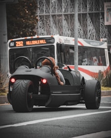 A sleek, black three-wheeled vehicle with red taillights is stopped at an urban intersection. A person wearing a furry coat is seated in the vehicle. In the background, a city bus with the destination 'Hillsdale Mall' is visible, indicating an urban setting. The scene captures a moment of contrast between modern, minimalistic vehicle design and the everyday public transportation system.
