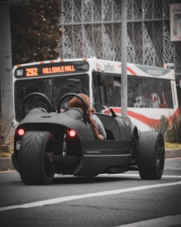 A sleek, black three-wheeled vehicle with red taillights is stopped at an urban intersection. A person wearing a furry coat is seated in the vehicle. In the background, a city bus with the destination 'Hillsdale Mall' is visible, indicating an urban setting. The scene captures a moment of contrast between modern, minimalistic vehicle design and the everyday public transportation system.