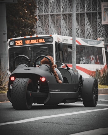 A sleek, black three-wheeled vehicle with red taillights is stopped at an urban intersection. A person wearing a furry coat is seated in the vehicle. In the background, a city bus with the destination 'Hillsdale Mall' is visible, indicating an urban setting. The scene captures a moment of contrast between modern, minimalistic vehicle design and the everyday public transportation system.