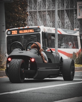 A sleek, black three-wheeled vehicle with red taillights is stopped at an urban intersection. A person wearing a furry coat is seated in the vehicle. In the background, a city bus with the destination 'Hillsdale Mall' is visible, indicating an urban setting. The scene captures a moment of contrast between modern, minimalistic vehicle design and the everyday public transportation system.