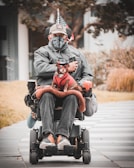 A person in a wheelchair, dressed in a grey jacket, cap, and face mask, holding a small dog that is dressed in a red outfit with glasses. The wheelchair is equipped with various accessories including a flag. The background includes a paved pathway, some grass, and trees.