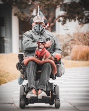 A person in a wheelchair, dressed in a grey jacket, cap, and face mask, holding a small dog that is dressed in a red outfit with glasses. The wheelchair is equipped with various accessories including a flag. The background includes a paved pathway, some grass, and trees.