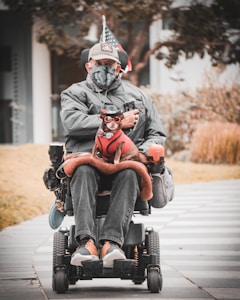 A person in a wheelchair, dressed in a grey jacket, cap, and face mask, holding a small dog that is dressed in a red outfit with glasses. The wheelchair is equipped with various accessories including a flag. The background includes a paved pathway, some grass, and trees.