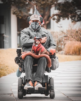 A person in a wheelchair, dressed in a grey jacket, cap, and face mask, holding a small dog that is dressed in a red outfit with glasses. The wheelchair is equipped with various accessories including a flag. The background includes a paved pathway, some grass, and trees.