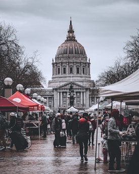 A bustling outdoor market scene with people walking and browsing stalls beneath overcast skies. The impressive architecture of a large domed building serves as a backdrop. Various tents line the sides, with vendors engaging with shoppers. Trees are visible, adding a natural element to the urban setting.