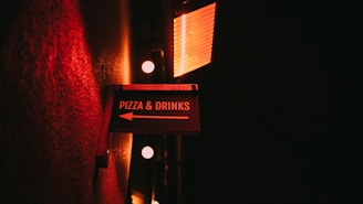A dimly lit sign directing towards pizza and drinks is mounted on a textured wall. The sign is illuminated by a warm red light, creating a cozy and inviting ambiance. The background is dark, enhancing the glow from the sign and nearby lights.
