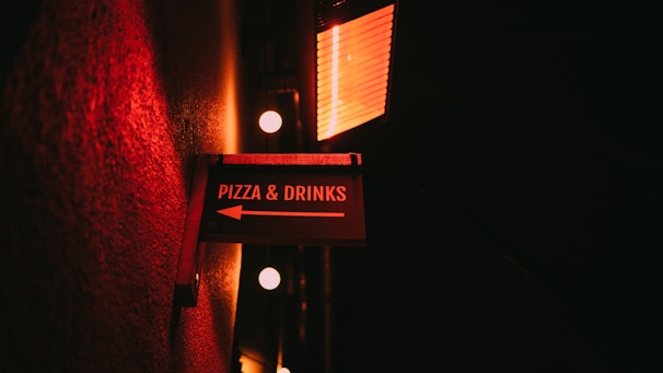 A dimly lit sign directing towards pizza and drinks is mounted on a textured wall. The sign is illuminated by a warm red light, creating a cozy and inviting ambiance. The background is dark, enhancing the glow from the sign and nearby lights.