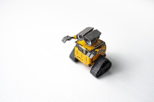 A small, toy-like yellow robot with large binocular-style eyes and track-like wheels is positioned on a smooth white surface. Its mechanical arm is slightly raised, and the design appears both endearing and functional.