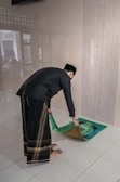 A person is unrolling or placing a prayer rug on a tiled floor. The individual is wearing traditional attire, which includes a black top and patterned pants. The setting appears to be indoors with a plain wall in the background.