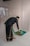 A person is unrolling or placing a prayer rug on a tiled floor. The individual is wearing traditional attire, which includes a black top and patterned pants. The setting appears to be indoors with a plain wall in the background.