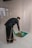 A person is unrolling or placing a prayer rug on a tiled floor. The individual is wearing traditional attire, which includes a black top and patterned pants. The setting appears to be indoors with a plain wall in the background.