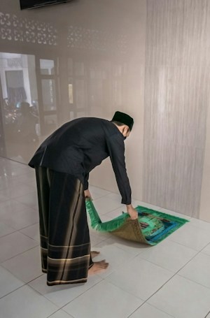 A person is unrolling or placing a prayer rug on a tiled floor. The individual is wearing traditional attire, which includes a black top and patterned pants. The setting appears to be indoors with a plain wall in the background.