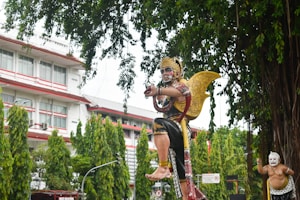 A large, ornate statue of a mythological figure with a golden crown and wings is prominently displayed. The figure is adorned in traditional attire with intricate patterns, and its expression conveys intensity. In the background, there is another smaller statue with a white face, also dressed traditionally. The scene is set outdoors with tall, lush green trees and a multi-story building with white walls and red accents in the background.