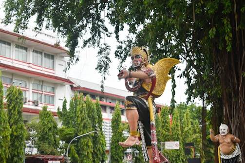 A large, ornate statue of a mythological figure with a golden crown and wings is prominently displayed. The figure is adorned in traditional attire with intricate patterns, and its expression conveys intensity. In the background, there is another smaller statue with a white face, also dressed traditionally. The scene is set outdoors with tall, lush green trees and a multi-story building with white walls and red accents in the background.