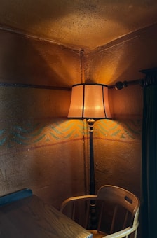 A warm, dimly lit corner of a room featuring a floor lamp with a beige lampshade casting light on textured walls. The walls have a decorative pattern with wavy lines. A wooden chair and part of a desk are positioned near the lamp. The overall ambiance is cozy and inviting.