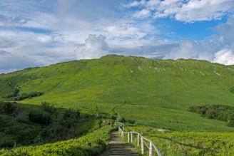 A lush green hillside with a wooden fence-lined path leading toward the top. The sky is partly cloudy with patches of bright blue visible. The landscape is expansive, offering a serene and open view.