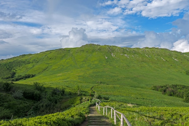 A lush green hillside with a wooden fence-lined path leading toward the top. The sky is partly cloudy with patches of bright blue visible. The landscape is expansive, offering a serene and open view.