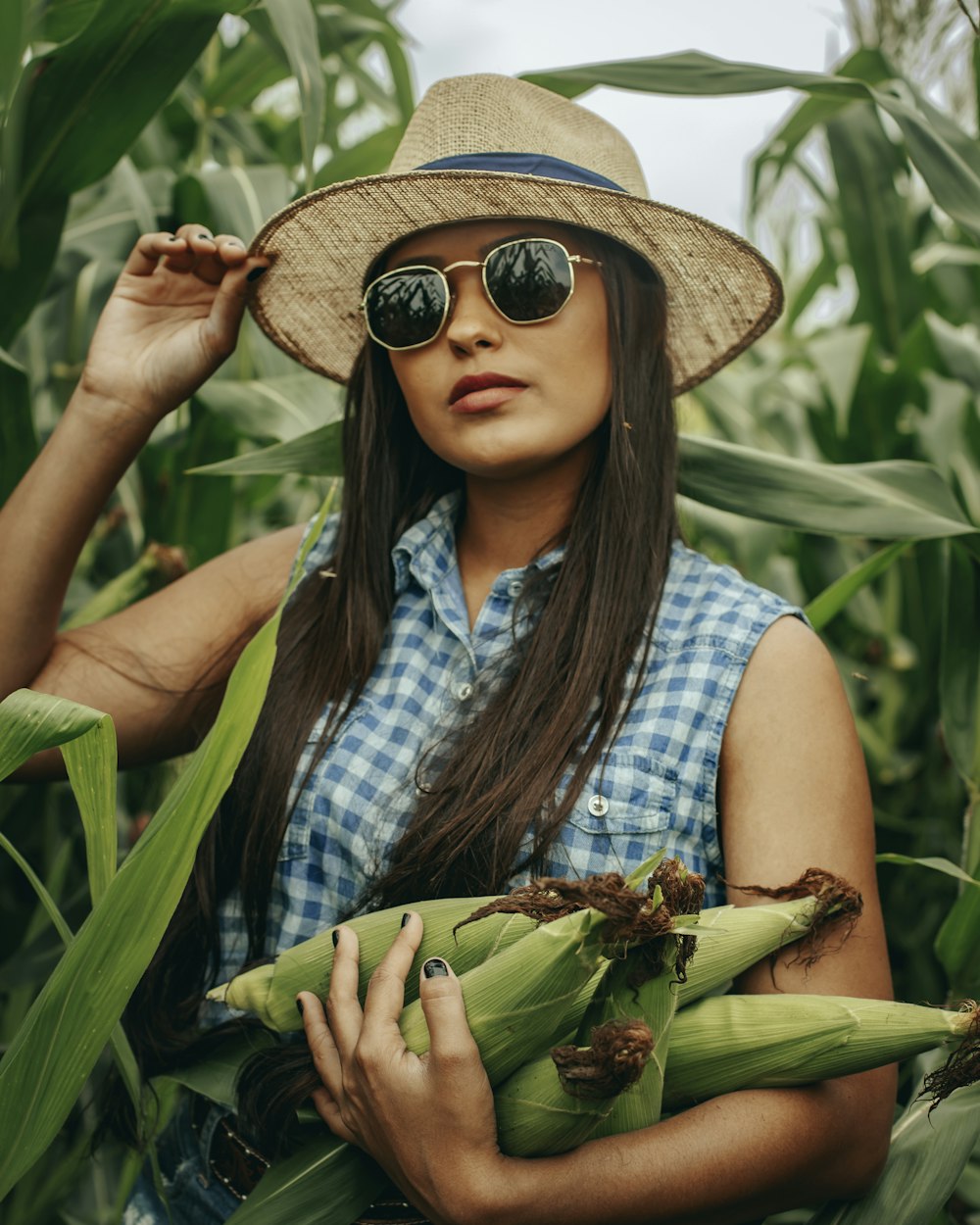 woman in blue and white plaid button up shirt and brown sun hat holding green banana