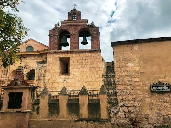 A historic church facade made of stone and brick featuring a bell tower with two large bronze bells and a cross at the top. The foreground shows a wall with pointed stone details and iron fencing. A street sign with 'UNA VIA' is visible on the right, and leafy branches frame the top left corner.