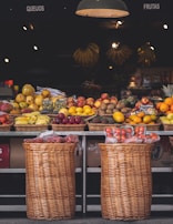 A market stall displays a variety of fresh fruits arranged in wicker baskets. The fruits include bananas, apples, mangos, oranges, and other colorful produce. Below, there are two large wicker baskets containing packaged goods. The setting is illuminated by warm overhead lights, creating a cozy atmosphere.