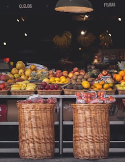 A market stall displays a variety of fresh fruits arranged in wicker baskets. The fruits include bananas, apples, mangos, oranges, and other colorful produce. Below, there are two large wicker baskets containing packaged goods. The setting is illuminated by warm overhead lights, creating a cozy atmosphere.