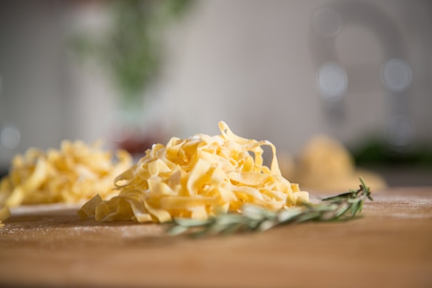 A close-up view of freshly made pasta, likely tagliatelle, on a wooden surface. The pasta is a vibrant yellow color and arranged in loose piles. In the foreground, there's a sprig of rosemary, adding a touch of green to the scene. The background is softly blurred, highlighting the pasta as the main focus.