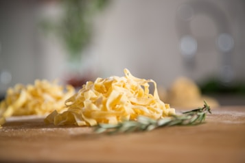A close-up view of freshly made pasta, likely tagliatelle, on a wooden surface. The pasta is a vibrant yellow color and arranged in loose piles. In the foreground, there's a sprig of rosemary, adding a touch of green to the scene. The background is softly blurred, highlighting the pasta as the main focus.