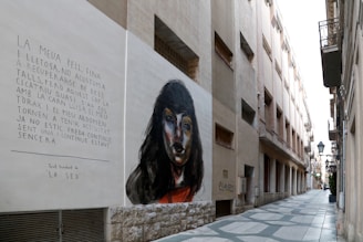 A narrow urban alley with buildings on both sides features a large mural on one wall. The mural depicts a woman's face with dark hair in a realistic style. Beside the painting, there is a text written in Catalan, which appears to be poetic in nature. The alleyway is paved with a geometric pattern, and several balconies and street lamps are visible along the buildings.
