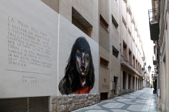 A narrow urban alley with buildings on both sides features a large mural on one wall. The mural depicts a woman's face with dark hair in a realistic style. Beside the painting, there is a text written in Catalan, which appears to be poetic in nature. The alleyway is paved with a geometric pattern, and several balconies and street lamps are visible along the buildings.