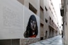 A narrow urban alley with buildings on both sides features a large mural on one wall. The mural depicts a woman's face with dark hair in a realistic style. Beside the painting, there is a text written in Catalan, which appears to be poetic in nature. The alleyway is paved with a geometric pattern, and several balconies and street lamps are visible along the buildings.