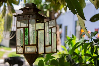 A rustic, stained-glass lantern hangs among green foliage outdoors, with a blurred background that includes a white building and various plants. The lantern features geometric patterns with green and white glass panes and appears aged with some visible cracks. Sunlight filters through the leaves and glass, creating a serene, nature-filled atmosphere.
