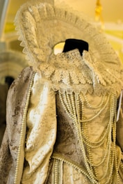An ornate historical costume featuring intricate lace detailing around the collar and pearls draped across the bodice. The fabric has an embroidered design with a mix of gold and cream colors, giving a vintage and opulent appearance.