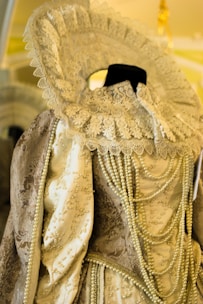An ornate historical costume featuring intricate lace detailing around the collar and pearls draped across the bodice. The fabric has an embroidered design with a mix of gold and cream colors, giving a vintage and opulent appearance.