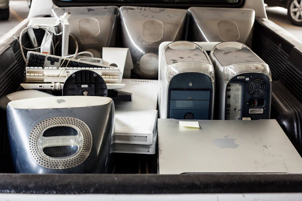 Several old desktop computer towers, including iMac G4s, old keyboards, a liquid cooling system, and other electronic items are placed in the back of a vehicle. The hardware appears used and dusty.