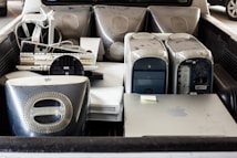 Several old desktop computer towers, including iMac G4s, old keyboards, a liquid cooling system, and other electronic items are placed in the back of a vehicle. The hardware appears used and dusty.