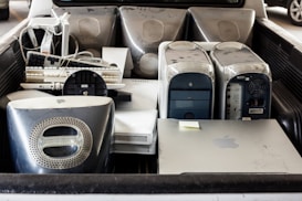 Several old desktop computer towers, including iMac G4s, old keyboards, a liquid cooling system, and other electronic items are placed in the back of a vehicle. The hardware appears used and dusty.