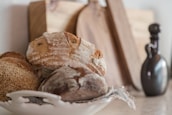A rustic setting featuring a white ceramic dish holding several artisan loaves of bread with crusty, textured surfaces. The background includes wooden cutting boards and a dark, curvy ceramic bottle, contributing to a cozy, kitchen atmosphere.