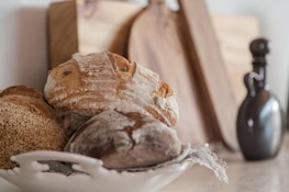 A rustic setting featuring a white ceramic dish holding several artisan loaves of bread with crusty, textured surfaces. The background includes wooden cutting boards and a dark, curvy ceramic bottle, contributing to a cozy, kitchen atmosphere.