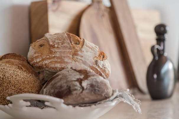 A rustic setting featuring a white ceramic dish holding several artisan loaves of bread with crusty, textured surfaces. The background includes wooden cutting boards and a dark, curvy ceramic bottle, contributing to a cozy, kitchen atmosphere.