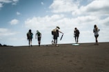 A group of people is walking up a sandy slope under a partly cloudy sky. Each person carries a backpack, and some have sandboards. The atmosphere is one of adventure and exploration.